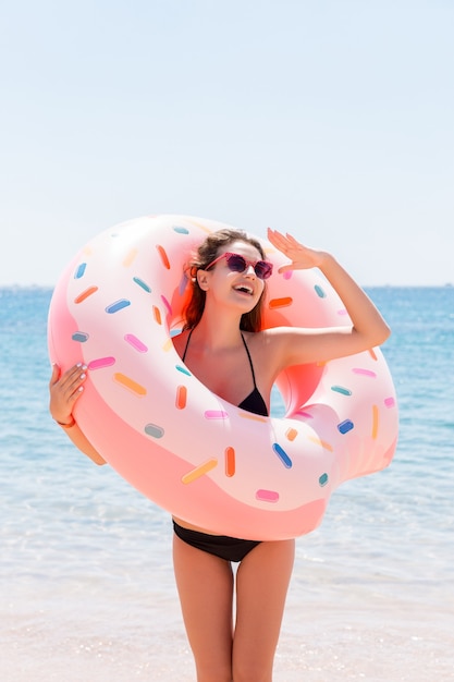 Disfrutando del bronceado y de las vacaciones. Retrato de una niña feliz mirando a través del anillo inflable permanecer en la playa del mar. Concepto de vacaciones de verano.