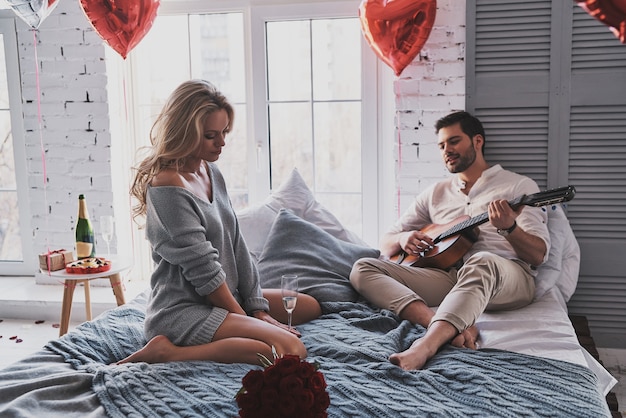 Disfrutando de buena música. Apuesto joven tocando la guitarra con su hermosa novia mientras celebra el Día de San Valentín en casa