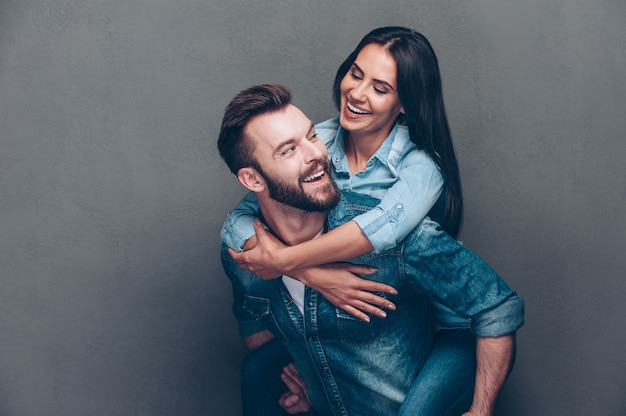 Disfrutando cada segundo juntos. Hombre joven hermoso que lleva a cuestas a la mujer hermosa y que sonríe mientras está de pie contra el fondo gris