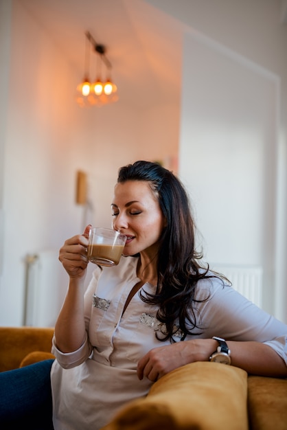 Disfrutando de un café en casa. Café de consumición de la morenita hermosa, sentándose en el sofá.