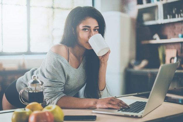 Disfrutando de un café recién hecho por la mañana. Hermosa joven mujer de raza mixta tomando café y mirando a la cámara mientras se inclina en el escritorio de la cocina en casa