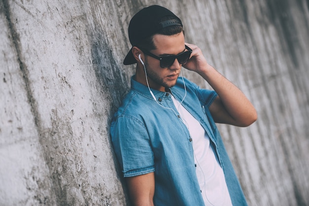 Disfrutando del estilo casual. Apuesto joven con gorra y gafas de sol escuchando música mientras usa auriculares y apoyado en la pared de hormigón