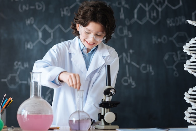 Disfrutando del experimento moderno. Alegre niño capaz sonriente de pie en el laboratorio y disfrutando de la lección de química mientras participa en el proyecto científico