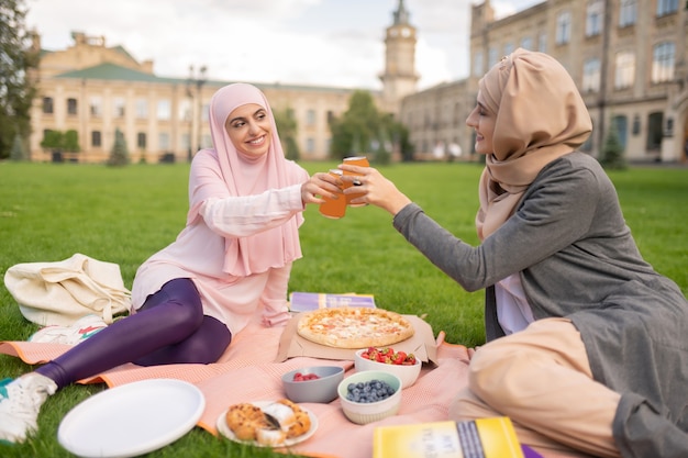 Disfrutando de la hora de la pizza. Estudiantes musulmanes alegres que disfrutan de la pizza al aire libre y beben bebidas gaseosas