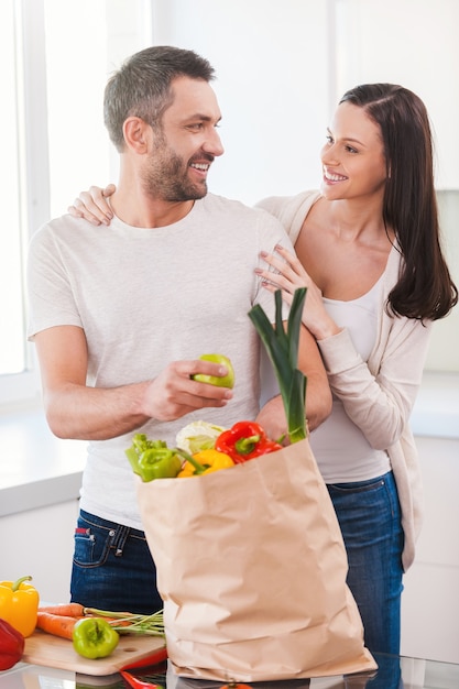 Disfrutando juntos de una vida feliz y saludable. Hermosa joven pareja desembalaje de la bolsa de la compra llena de verduras frescas y sonriendo mientras está de pie en la cocina juntos