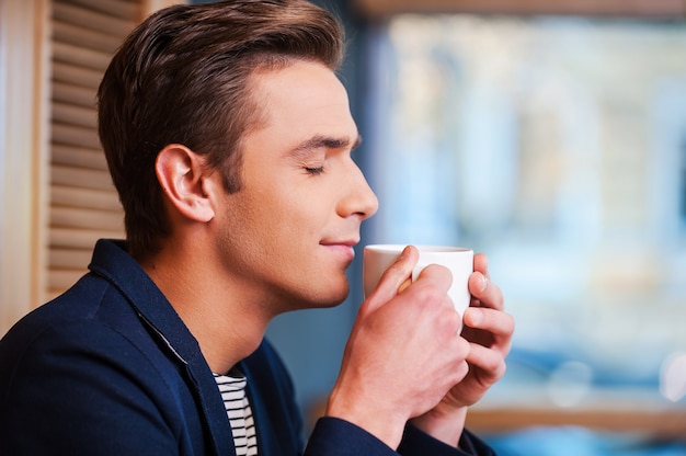 Disfrutando del mejor café de la ciudad. Vista lateral del apuesto joven manteniendo los ojos cerrados y sonriendo mientras huele una taza de café recién hecho en la cafetería