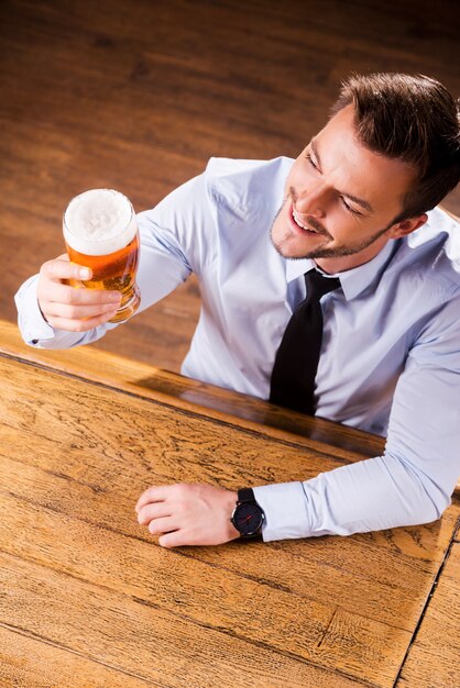 Disfrutando de la mejor cerveza de la ciudad. Vista superior del apuesto joven en camisa y corbata examinando el vaso con cerveza y sonriendo mientras está sentado en la barra del bar