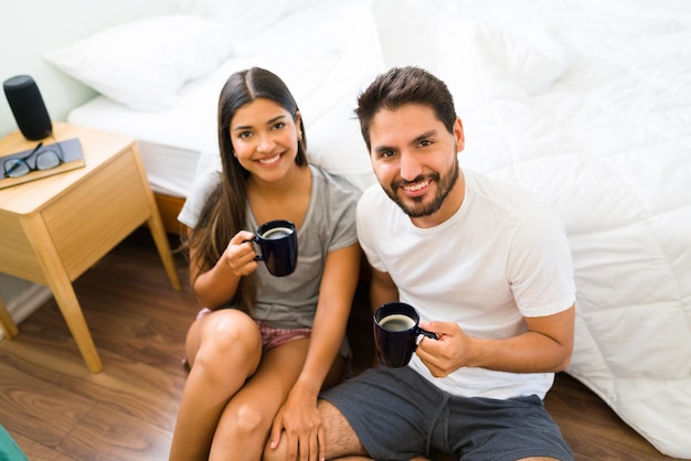 Disfrutando de un momento de relax en la mañana. Atractiva pareja en pijama sonriendo y haciendo contacto visual mientras bebe una taza de café caliente en el dormitorio