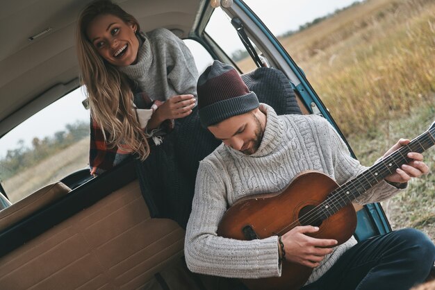 Disfrutando de momentos felices. Apuesto joven tocando la guitarra para su hermosa novia mientras está sentado en una mini furgoneta de estilo retro