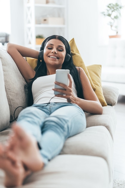 Disfrutando de la música. Mujer hermosa joven escuchando música y sonriendo mientras está acostado en el sofá en casa