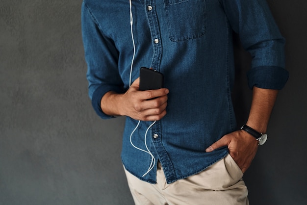 Disfrutando de la música. Primer plano de un joven sosteniendo un teléfono inteligente y manteniendo la mano en el bolsillo mientras está de pie contra el fondo gris