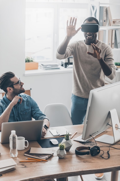 ¡Disfrutando de la nueva realidad! Dos hombres jóvenes exitosos en ropa casual trabajando en una oficina creativa mientras uno de ellos está parado en un casco de realidad virtual