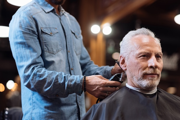 Disfrutando del servicio. Ángulo bajo de agradable hombre barbudo senior sentado mientras peluquero recorta su cabello en la barbería.