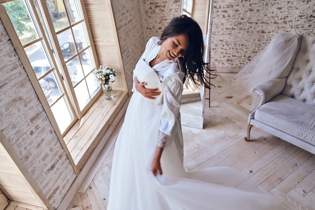 Disfrutando de su día especial. Vista superior de la atractiva joven en bata de baño de seda probándose su vestido de novia y sonriendo mientras está de pie junto a la ventana