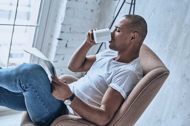 Disfrutando de su fin de semana. Vista superior del apuesto joven africano leyendo el periódico y tomando café