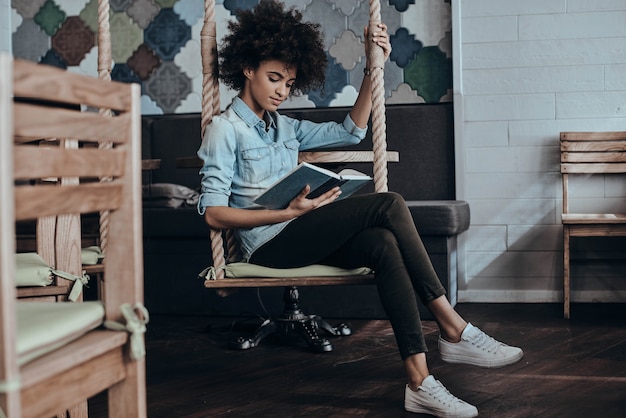 Disfrutando de su libro favorito. Hermosa joven africana leyendo un libro mientras está sentado en el columpio en la cafetería o en la oficina creativa