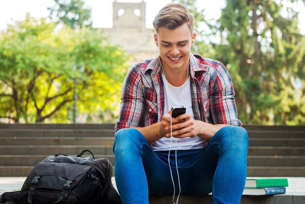 Disfrutando de su música favorita. Estudiante masculino feliz escuchando el reproductor MP3 y sonriendo mientras está sentado en la escalera al aire libre con libros y mochila cerca de él