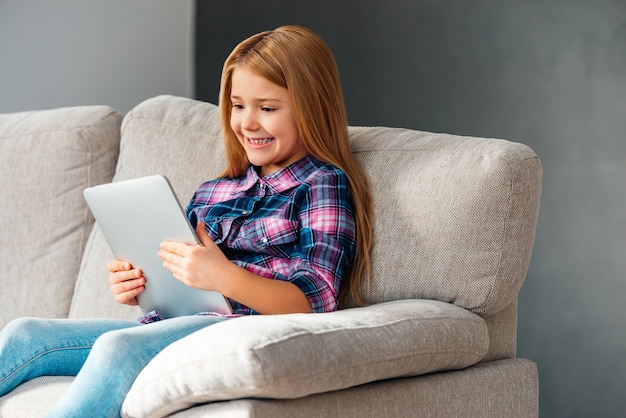 Disfrutando de su nueva tableta digital. Niña alegre mirando su panel táctil con una sonrisa mientras está sentado en el sofá en casa