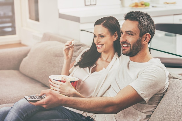 Disfrutando de su programa favorito. Hermosa joven pareja amorosa uniéndose entre sí y comiendo palomitas de maíz mientras está sentado en el sofá y ve la televisión