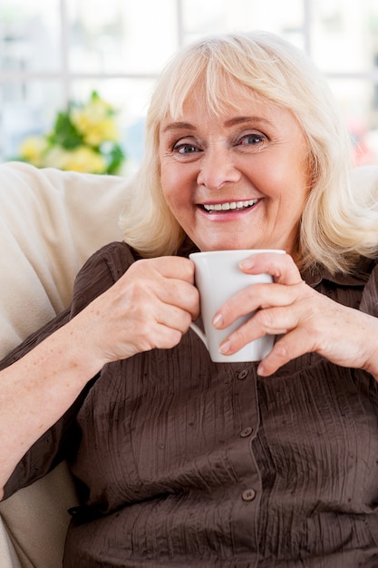 Disfrutando de un té caliente. Mujer mayor sosteniendo la taza y sonriendo mientras está sentado en la silla