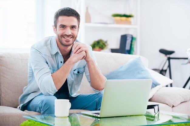 Disfrutando del tiempo en casa. Apuesto joven mirando a cámara y sonriendo mientras está sentado en el sofá en casa con un portátil cerca de él