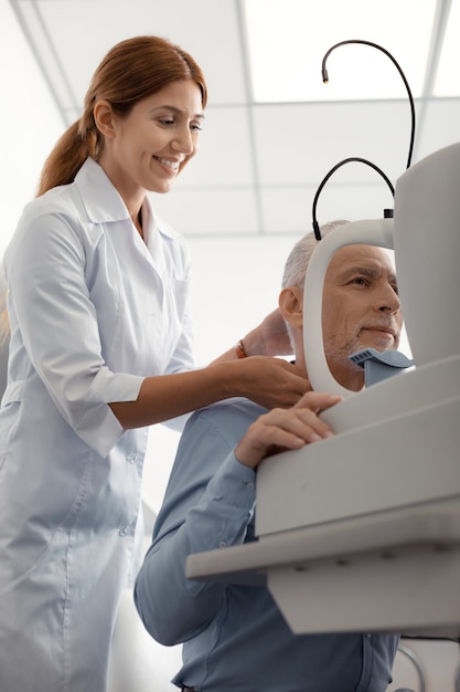 Disfrutando del trabajo. Agradable especialista en ojos radiantes disfrutando del tiempo en el trabajo mientras consulta a un anciano