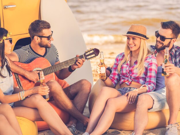 Disfrutando del verano con amigos. Grupo de jóvenes felices divirtiéndose juntos mientras está sentado en la playa cerca de su minivan