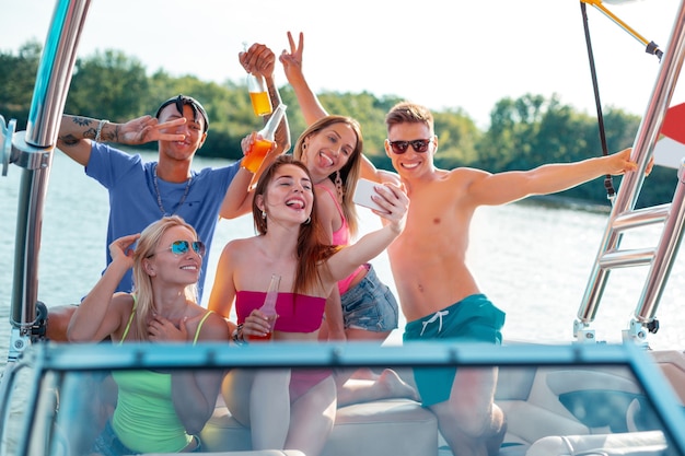 Disfrutando de la vida. Grupo de amigos alegres bebiendo refrescos y tomando selfies en un yate mirando directamente a la cámara