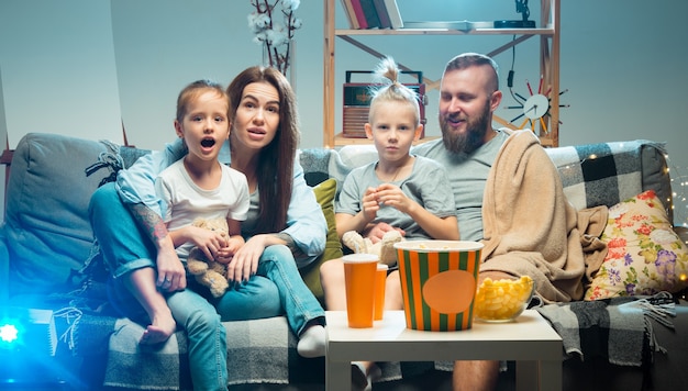 Disfrutar. Familia feliz viendo proyector, TV, películas con palomitas de maíz y bebidas por la noche en casa. Madre, padre e hijos pasando tiempo juntos. Confort en el hogar, tecnologías modernas, concepto de emociones.