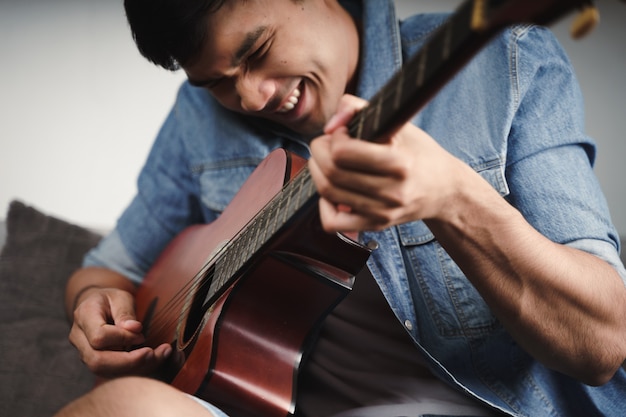 Disfrute de un apuesto hombre asiático practicando o tocando la guitarra en el sofá de la sala de estar