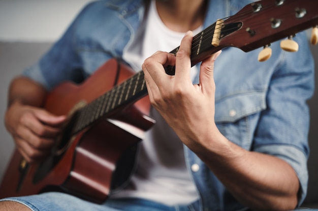 Disfrute de un apuesto hombre asiático practicando o tocando la guitarra en el sofá de la sala de estar