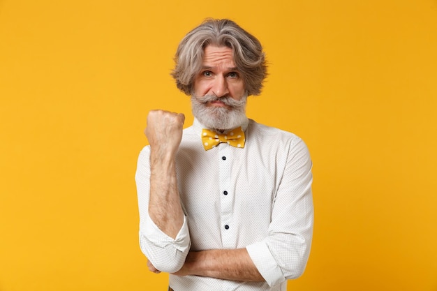 Disgustado anciano de pelo gris bigote barbudo hombre con camisa blanca, pajarita posando aislado sobre fondo amarillo naranja, retrato de estudio. Concepto de estilo de vida de las personas. Simulacros de espacio de copia. Puño apretado.