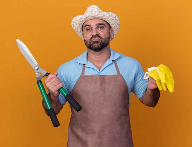 Disgustado jardinero varón caucásico adulto vistiendo sombrero de jardinería sosteniendo guantes y tijeras de jardinería