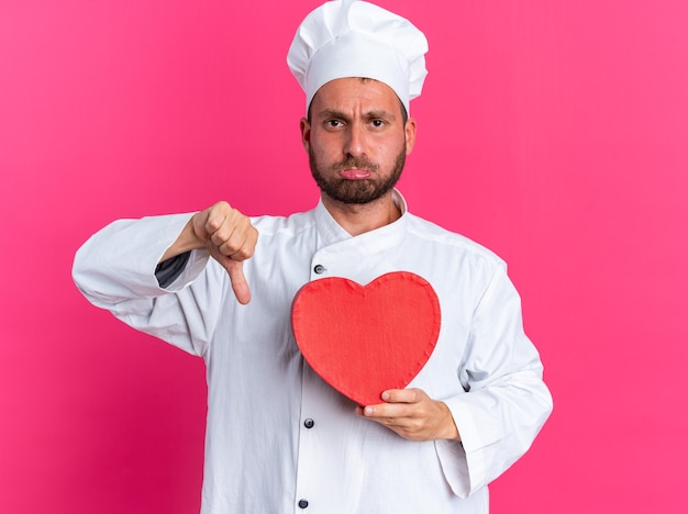 Disgustado joven cocinero de sexo masculino caucásico en uniforme de chef y gorra sosteniendo en forma de corazón mostrando el pulgar hacia abajo