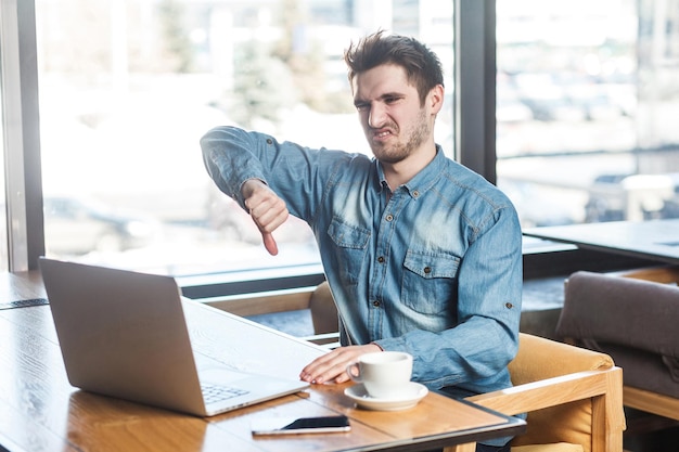 ¡Disgusto! Retrato de vista lateral de crítica negativa barbudo joven autónomo en camisa de jeans está sentado en la cafetería y haciendo videollamadas en la computadora portátil y mostrando el pulgar hacia abajo a su compañero. interior, interior