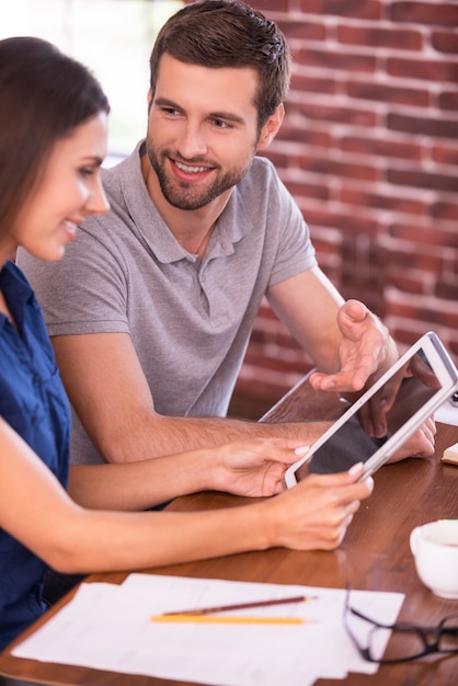 Diskussion über ein neues Geschäftsprojekt. Seitenansicht eines fröhlichen jungen Mannes und einer Frau, die am Tisch sitzen und reden, während der Mann auf ein digitales Tablet zeigt und lächelt