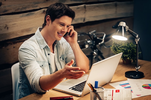 Diskussion über neues Projekt. Fröhlicher junger Mann, der am Handy spricht und den Laptop ansieht, während er an seinem Arbeitsplatz sitzt