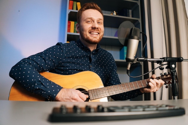 Dispara desde abajo de un guitarrista riendo, un hombre toca en una grabación de guitarra acústica, canta una canción usando un soporte de micrófono profesional en el estudio de casa