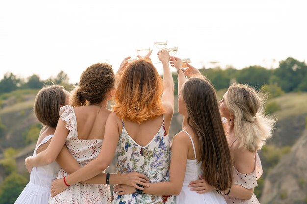 Dispara desde atrás. compañía de hermosas amigas divirtiéndose, animando y bebiendo vino, y disfruta de un picnic en las colinas.