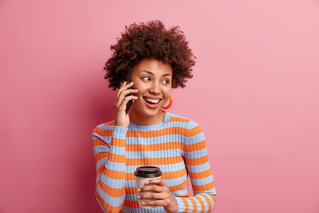 Disparo aislado de una mujer guapa con cabello afro que se ve felizmente lejos mantiene el teléfono inteligente cerca de la oreja, bebidas, café para llevar, usa un jersey de rayas casual aislado sobre una pared rosa