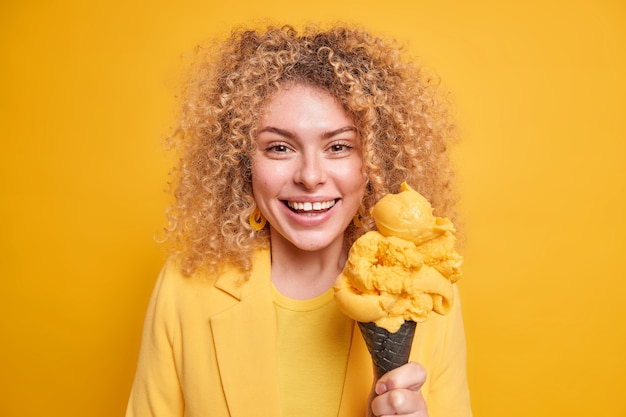 Disparo aislado de sonrisas positivas de adolescente de pelo rizado disfruta con alegría de un agradable día de verano y tiene un sabroso helado en gofre negro que no mantiene la dieta aislada sobre la pared amarilla. Concepto de estilo de vida