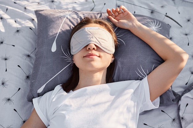 Disparo al aire libre de una joven morena vestida con camiseta blanca de estilo casual y antifaz para dormir acostado en la cama y durmiendo, disfrutando del aire fresco y el sol.