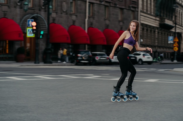 Disparo al aire libre de una mujer delgada activa vestida con top corto y patines en leggings a través de la ciudad disfruta de un excelente ejercicio físico quema calorías obtiene energía alivia el estrés aprende nuevas habilidades