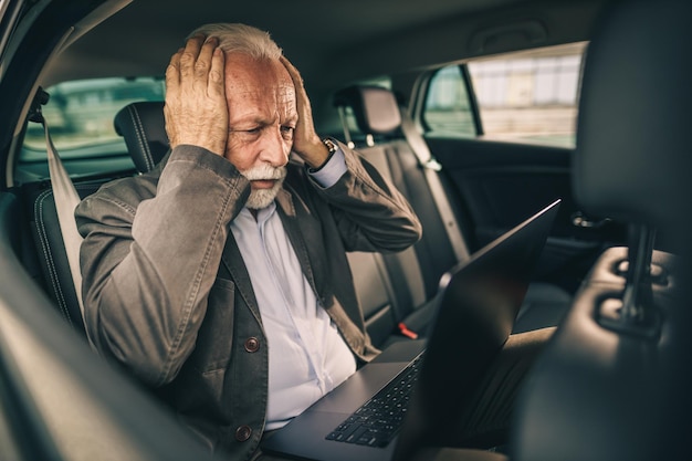 Disparo de un anciano estresado que usa una computadora portátil mientras está sentado en el asiento trasero de un automóvil durante su viaje de negocios.