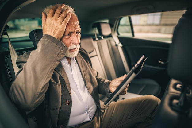 Disparo de un anciano preocupado que usa una tableta digital mientras se sienta en el asiento trasero de un automóvil durante su viaje de negocios.