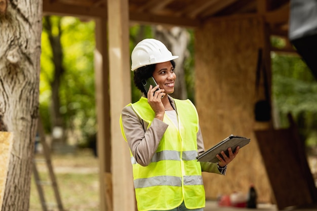 Disparo de una arquitecta africana usando un teléfono inteligente y una tableta digital mientras revisa el sitio de construcción de una nueva casa de madera. Lleva ropa de trabajo protectora y casco blanco.