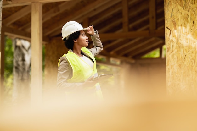 Disparo de una arquitecta afroamericana revisando el sitio de construcción de una nueva casa de madera. Lleva ropa de trabajo protectora y casco blanco.