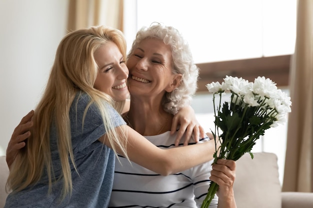 Disparo en la cabeza alegre hija adulta abrazando a la anciana madre diciendo felicitaciones con cumpleaños u ocasión especial presentando flores de campo Agradecida anciana abrazando a su nieta