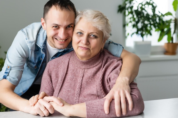 Disparó a la cabeza a un joven barbudo feliz abrazando a una hermosa y sonriente madre de mediana edad con anteojos, relajándose juntos en un cómodo sofá en la sala de estar, disfrutando de una conversación divertida o chismeando.