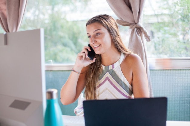 Disparo a la cabeza joven mujer feliz sentada en un escritorio con una llamada con su teléfono móvil, trabajando en la computadora en casa